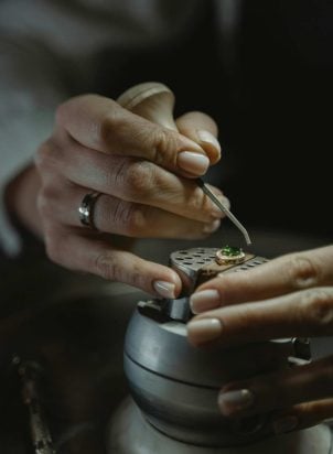 A jewelry artisan is setting small diamonds in a halo setting around an emerald.